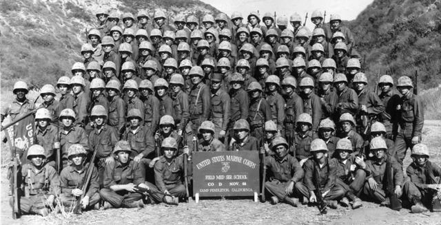 Field Medical Service School, Camp Pendleton, California, 1966. Russell is seated cross legged, second to the right of the sign, holding a rifle. The sign reads November 1964; the family dates the photo to 1966.